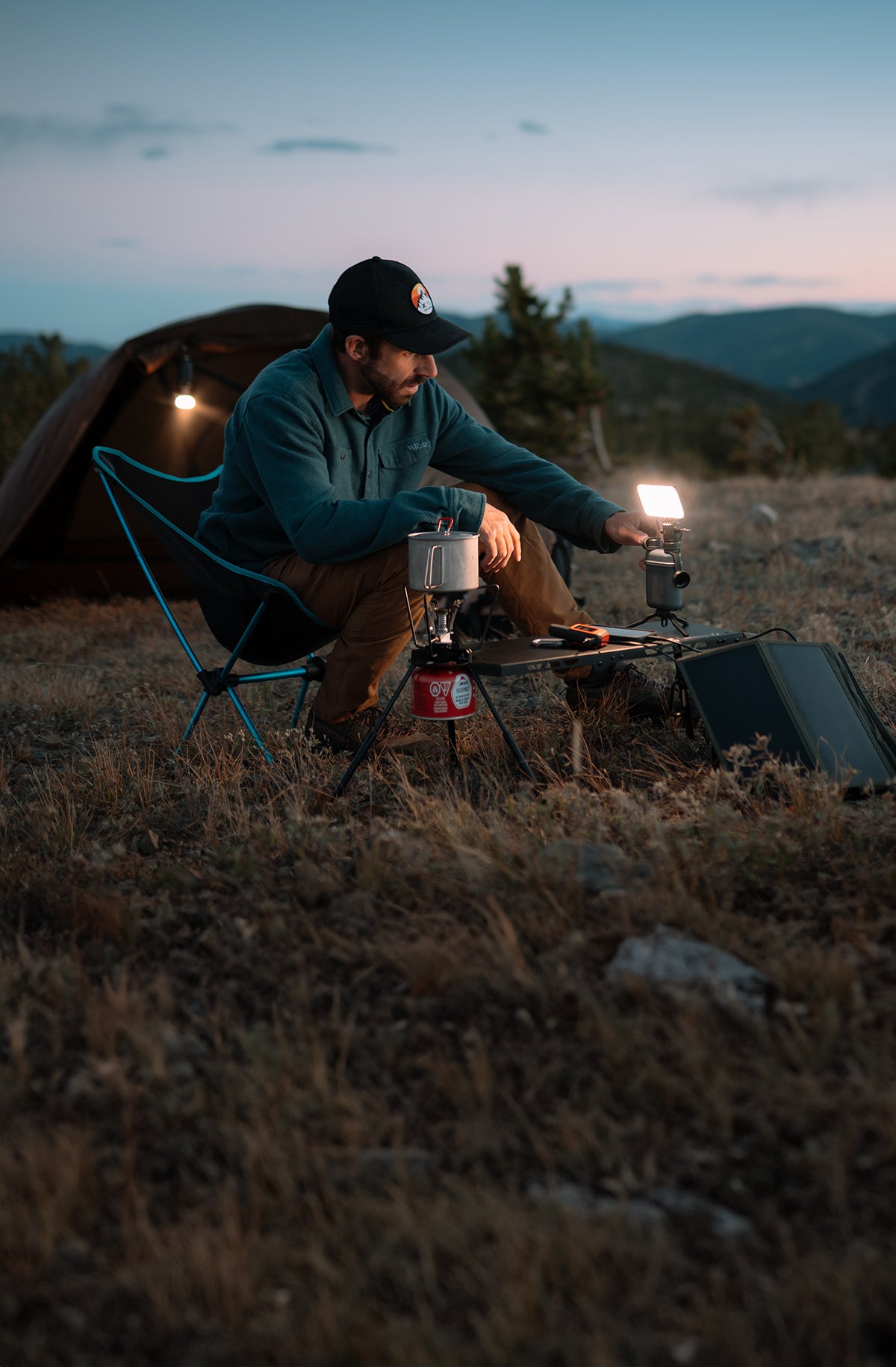 Man camping in a field with a lantern and cooking equipment at dusk.