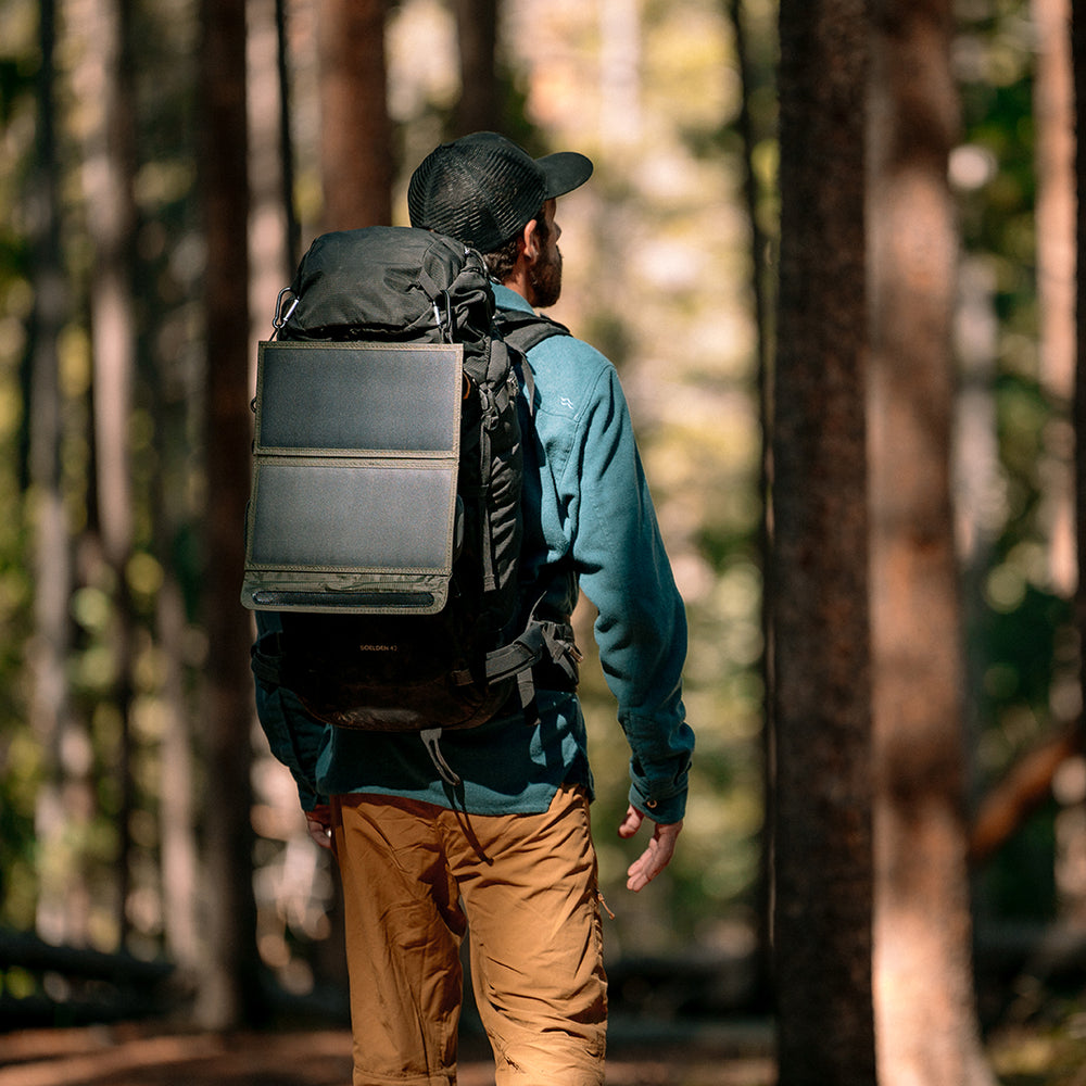 Person with a backpack that has a 2 panel solar charger attached to it and walking through a forest