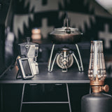Camp kitchen setup with a stove, kettle, lantern and CO monitor on a black surface.