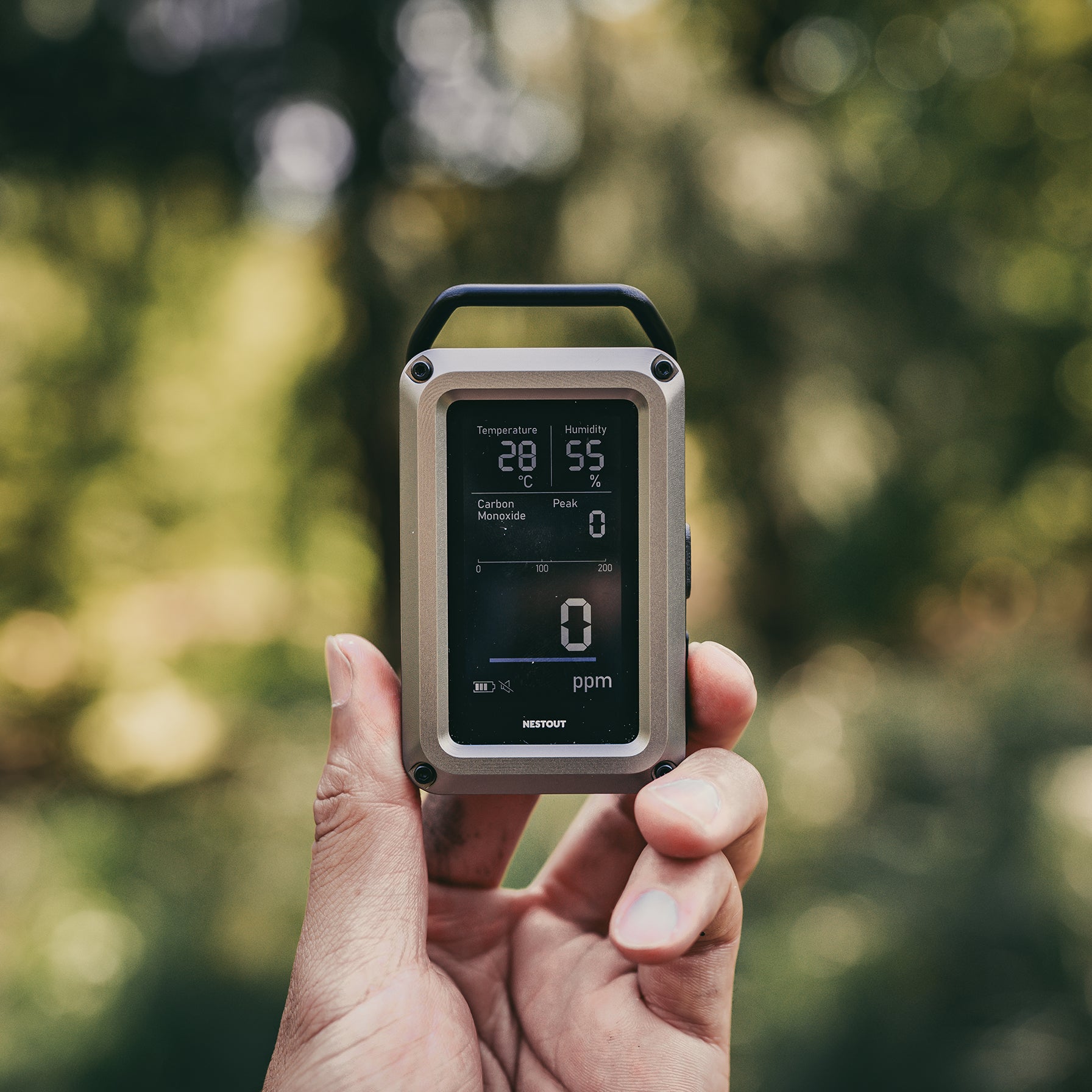 Hand holding a CO monitor with a screen displaying readings against a blurred green outdoor background