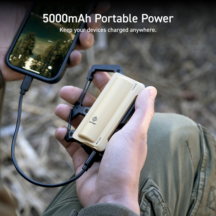 Text overlay reads: 5000mAh portable power: keep your devices charged anywhere | zoomed in image of a man holding a phone in his left hand and the beige nestout carabiner in his right hand while the phone charges 