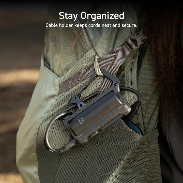 Text overlay in top middle center reads: Stay organized: cable holder keeps cords neat and secure. | image features a black carabiner power bank clipped to a two-tone sage green backpack worn by a brunette with long hair