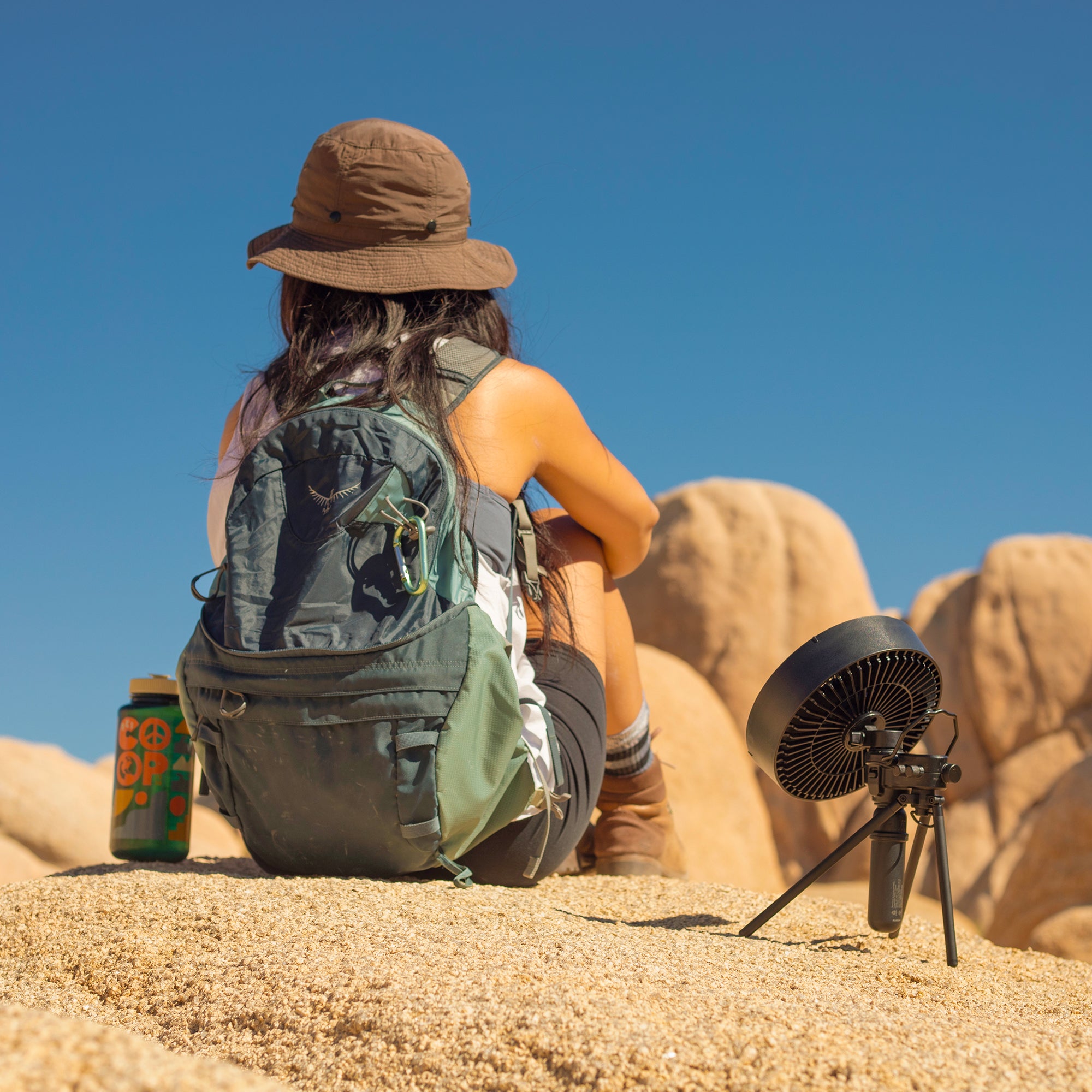 Image of female hiker sitting on rocks in joshua tree with black fan with black tripod attached and 5k battery attached