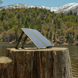 image of the nestout olive green 2 solar panel unfolded taking in sun resting on a tree stump with the lake and mountains in the background in Jenks Lake
