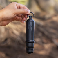 A hand holding a black cylindrical outdoor battery with a metal loop at the top, set against a blurred natural outdoor background.