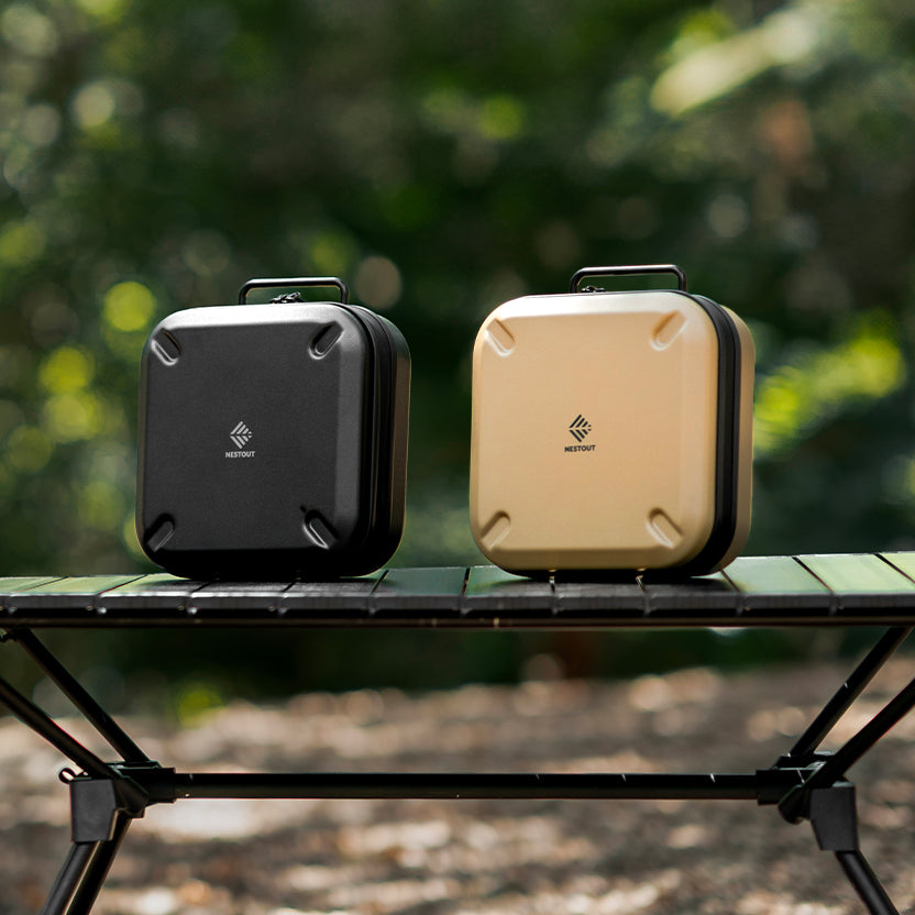 Two hard-shell square cases, one black and one beige, sitting on a folding table outdoors with a blurred green forest background.