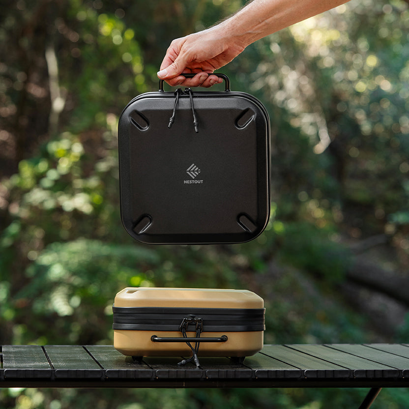 A hand holds a black rectangular carrying case above a tan and black case resting on a table outdoors, with a blurred forest background. The black case has a visible NESTOUT logo.