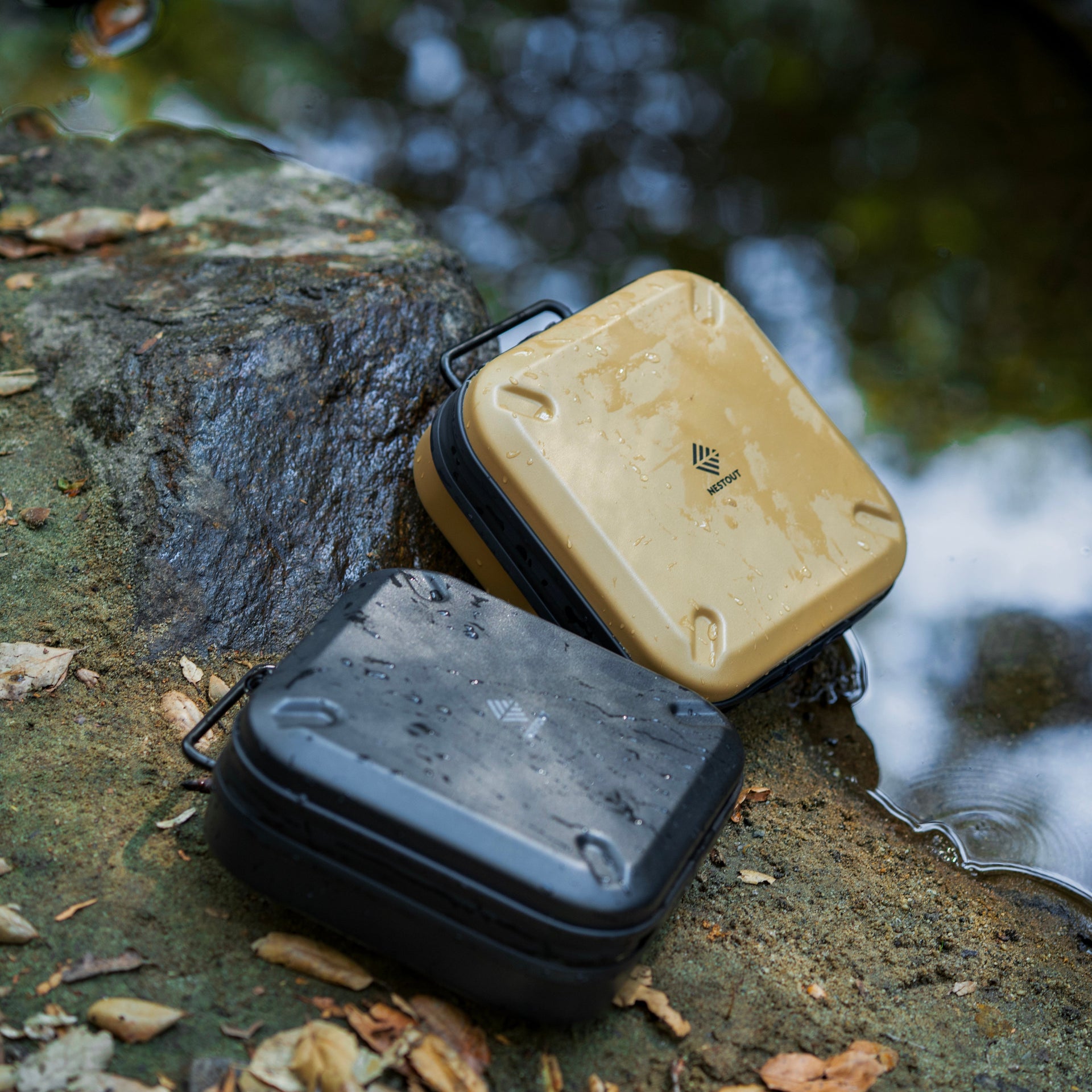 Two small electronic devices, one black and one beige, on a rocky surface near water.