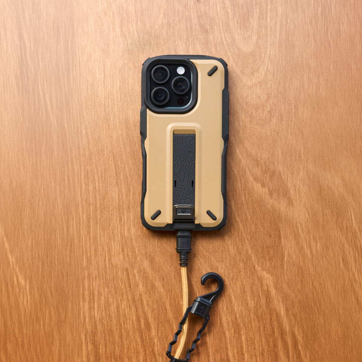 A rugged tan and black phone case with a built-in kickstand, shown from the back on a wooden surface. A black and tan wrist strap with a hook clasp is attached below the case.