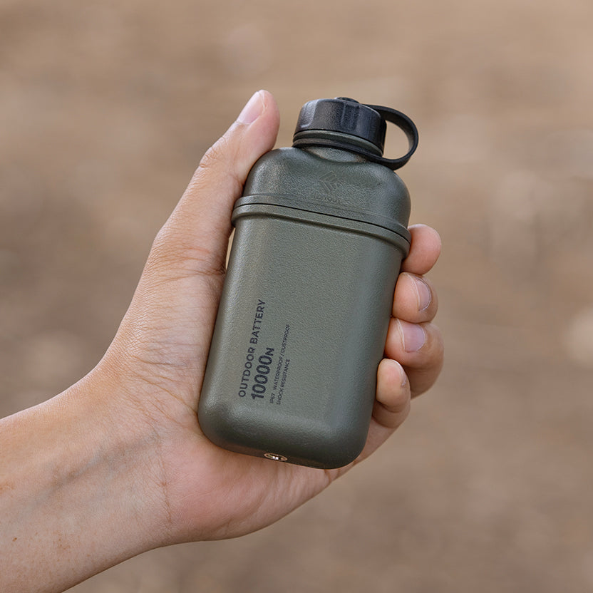 A hand holding an olive green outdoor battery with a black cap and loop, featuring the text 'OUTDOOR BATTERY 10000mAh' on the side, against a blurred brown background.