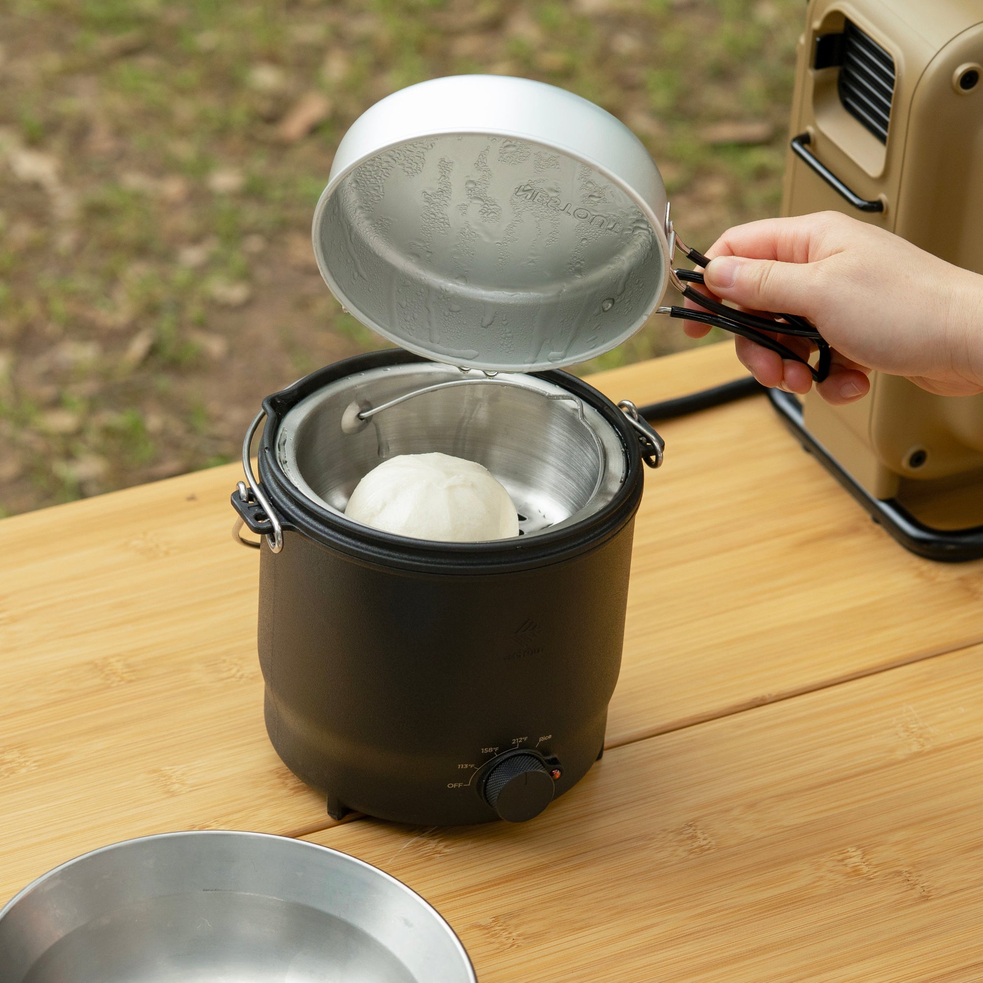 Person using a portable electric stove to steam dumplings on a wooden table outdoors.