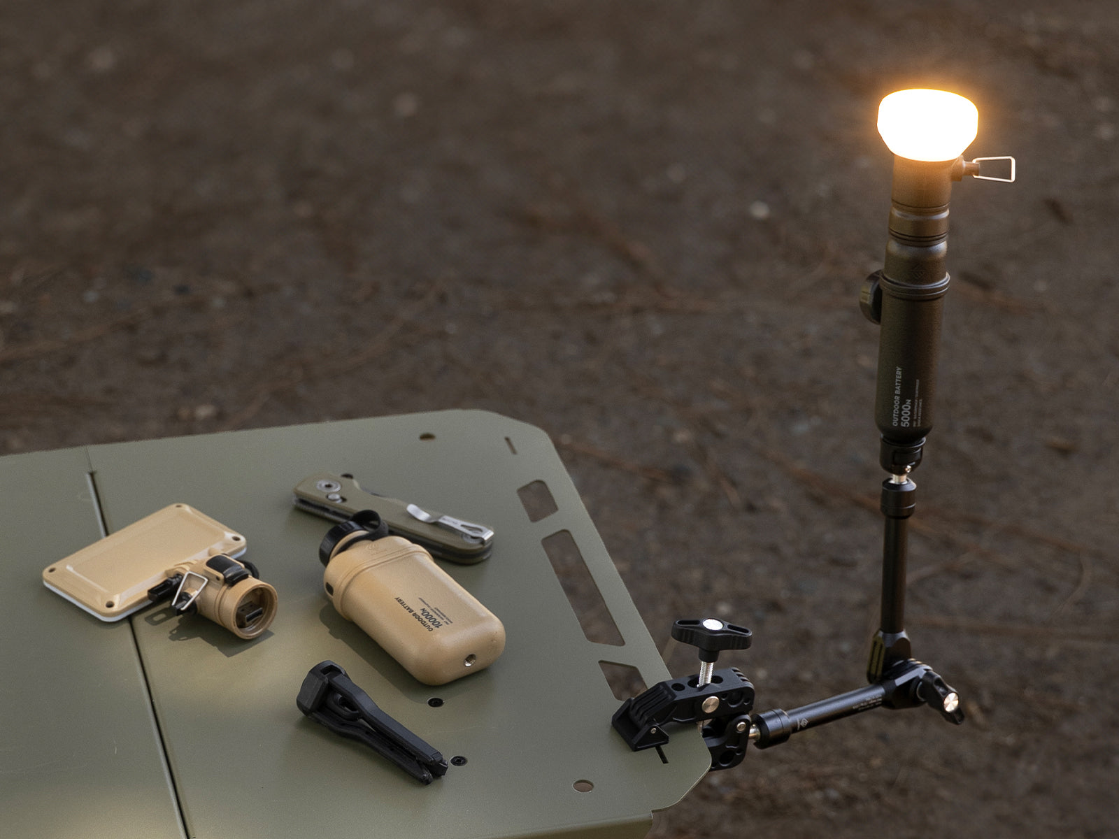 black clamp attached to table while the arm has a black 5 power bank attached with a lamp attached to that along with several accessories on a table