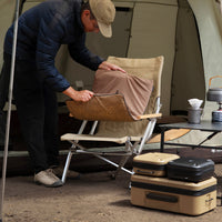 A man in a navy puffer jacket places a beige heating pad onto a camping chair at the entrance of a tent, with a folding table and hard cases set up nearby.
