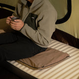 A person in a fleece jacket holds a mug while sitting on a beige heating pad placed on a sleeping pad inside a tent.