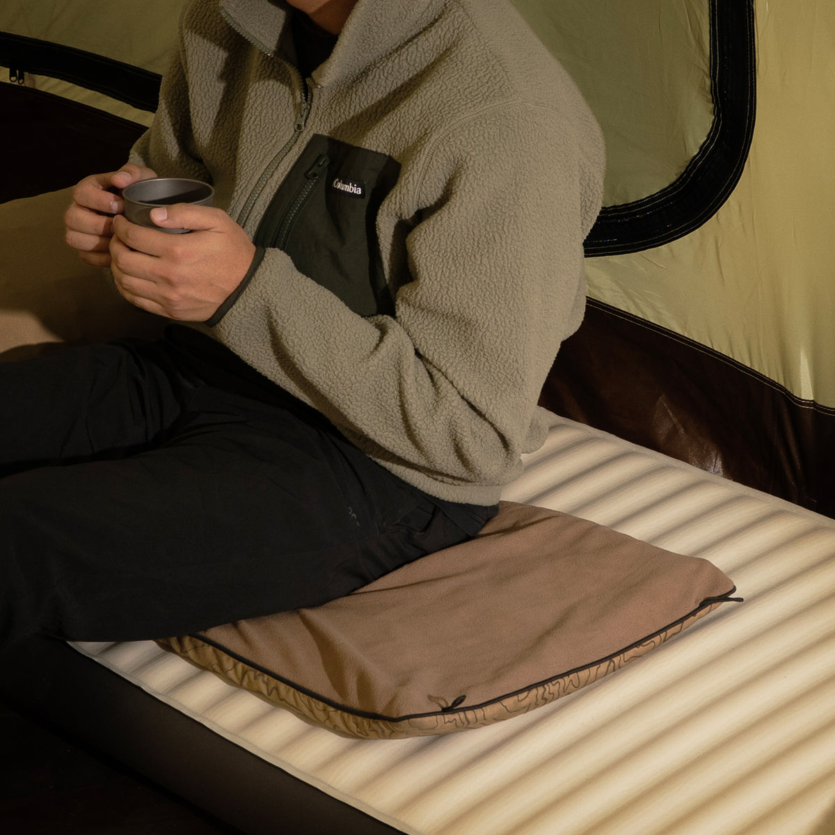 A person in a fleece jacket holds a mug while sitting on a beige heating pad placed on a sleeping pad inside a tent.