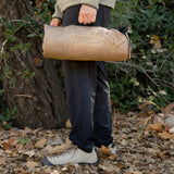 A person carries a rolled-up heating pad with a topographic contour pattern by its handle, standing outdoors on a path covered in fallen leaves.