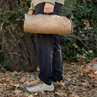 A person carries a rolled-up heating pad with a topographic contour pattern by its handle, standing outdoors on a path covered in fallen leaves.