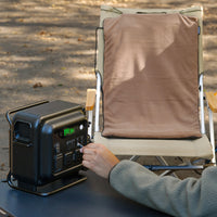 A person plugs a beige heating pad into a black portable power station on an outdoor table, with the pad draped over a camping chair behind it.