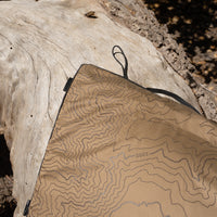 A close-up of a beige heating pad with a topographic contour pattern and black trim, laid over a log outdoors.