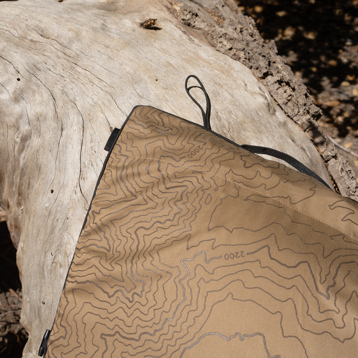 A close-up of a beige heating pad with a topographic contour pattern and black trim, laid over a log outdoors.