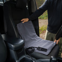 A person places a gray heating pad onto a car's front seat, with the controller and cable resting on the seat.