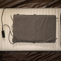 A gray heating pad laid flat on a sleeping pad inside a tent, with its controller and cable beside it.