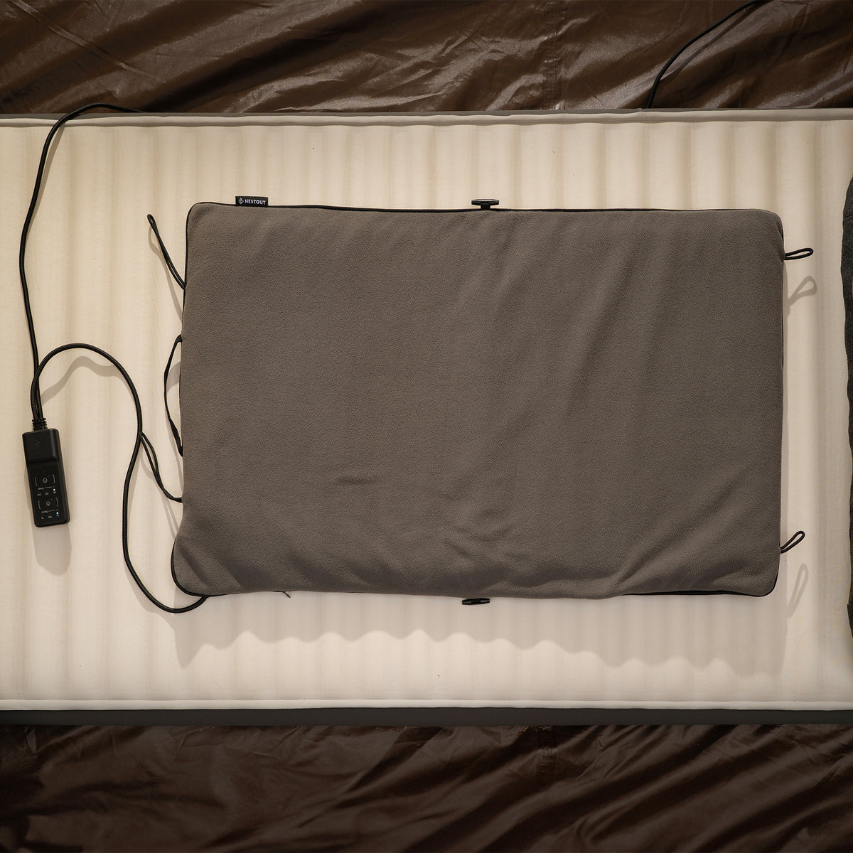 A gray heating pad laid flat on a sleeping pad inside a tent, with its controller and cable beside it.