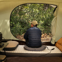 A man sits on a gray heating pad inside a tent, looking out the open door toward the trees.