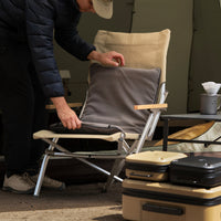 A man in a navy puffer jacket places a gray heating pad onto a camping chair at the entrance of a tent, with a folding table and hard cases set up nearby.