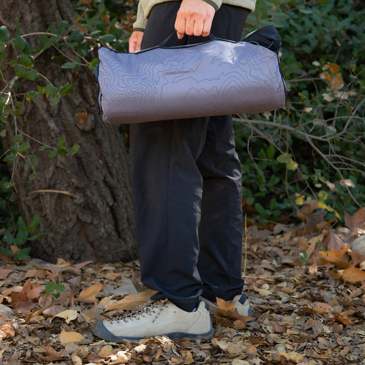A person carries a rolled-up gray heating pad with a topographic contour pattern by its handle, standing outdoors on a path covered in fallen leaves.