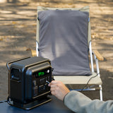 A person plugs a gray heating pad into a black portable power station on an outdoor table, with the pad draped over a camping chair behind it.