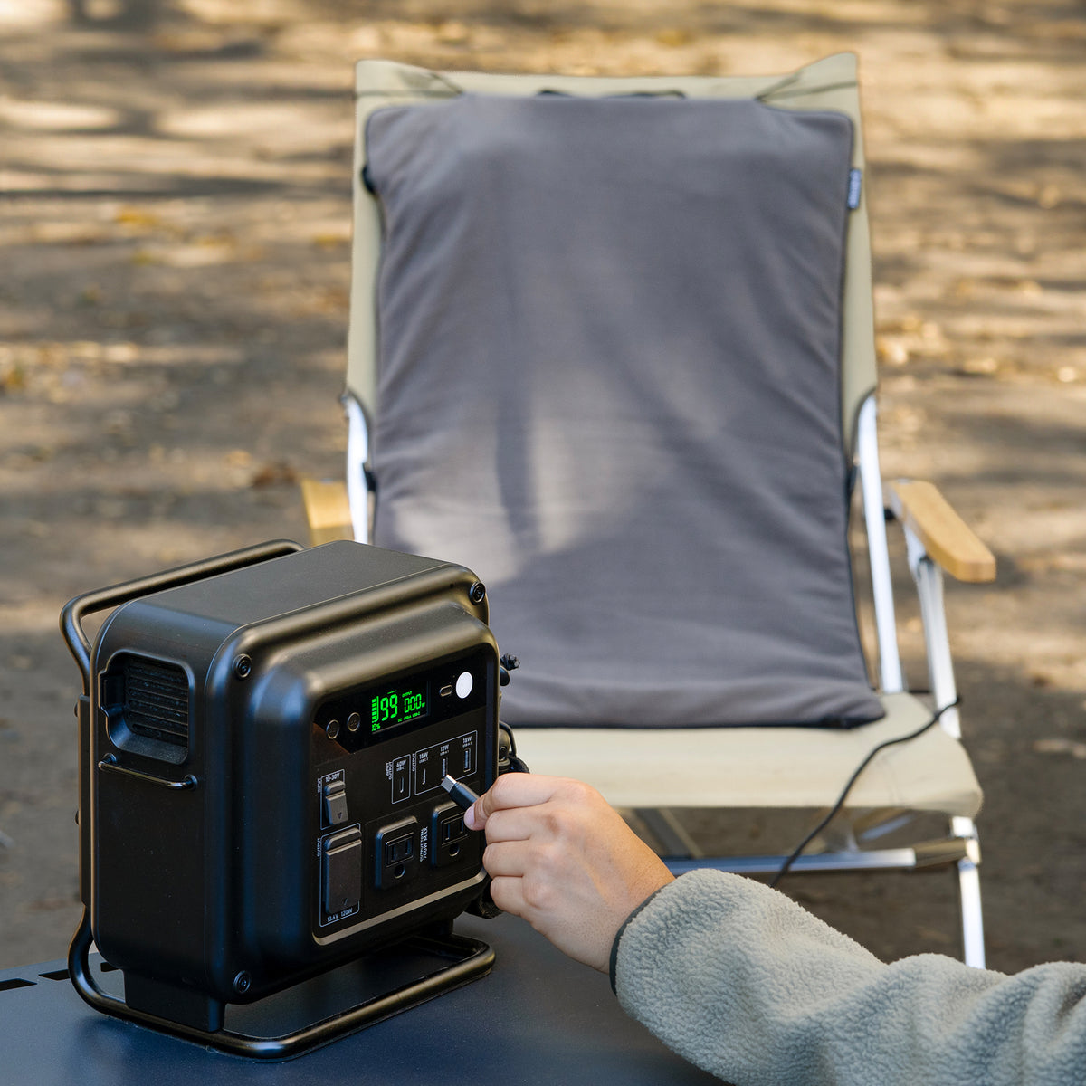 A person plugs a gray heating pad into a black portable power station on an outdoor table, with the pad draped over a camping chair behind it.