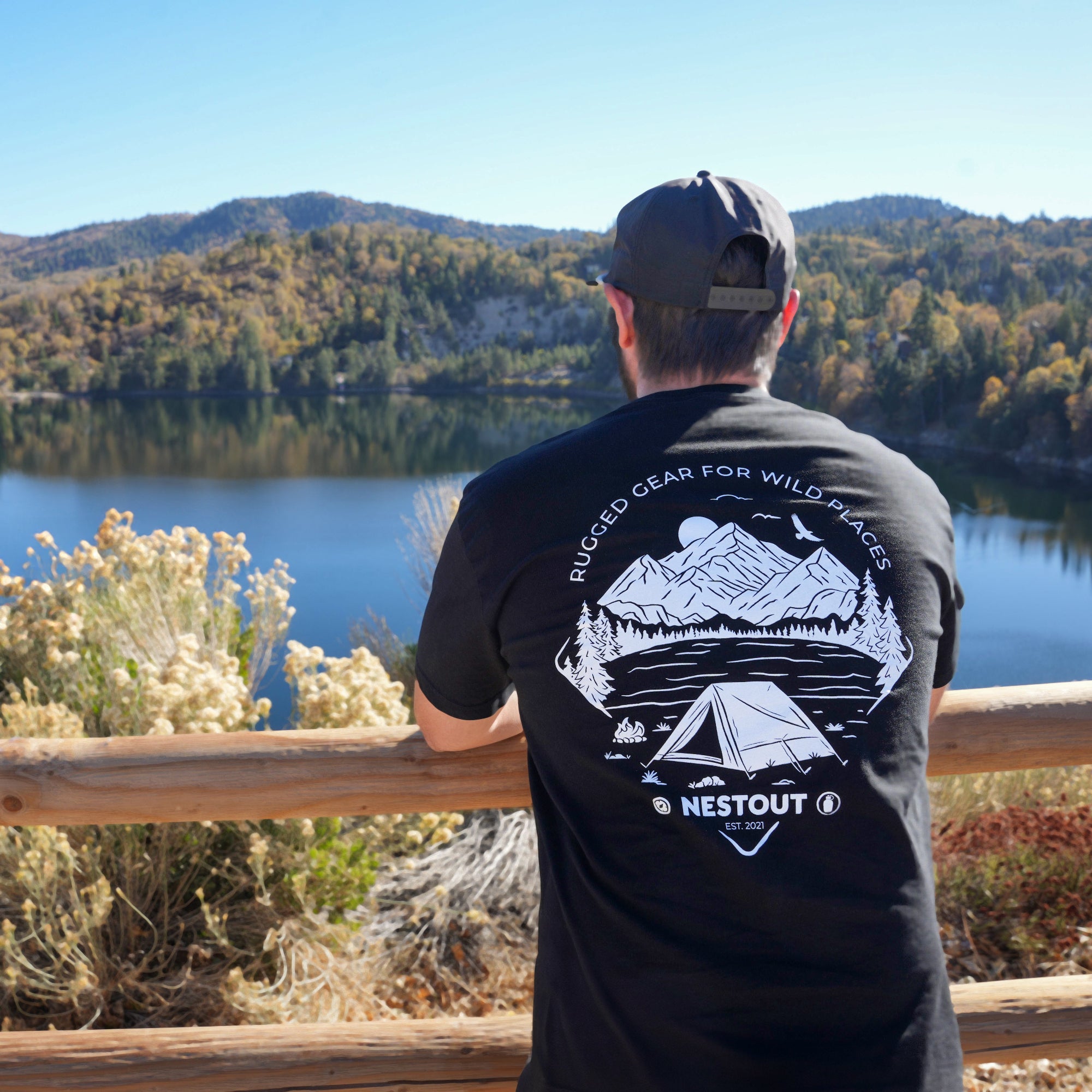 Person wearing a black t-shirt with a mountain and tent design, standing by a lake with mountains in the background.
