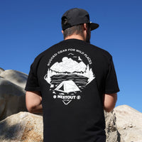 Person wearing a black t-shirt with a mountain and tent design, standing on rocks with a clear blue sky.