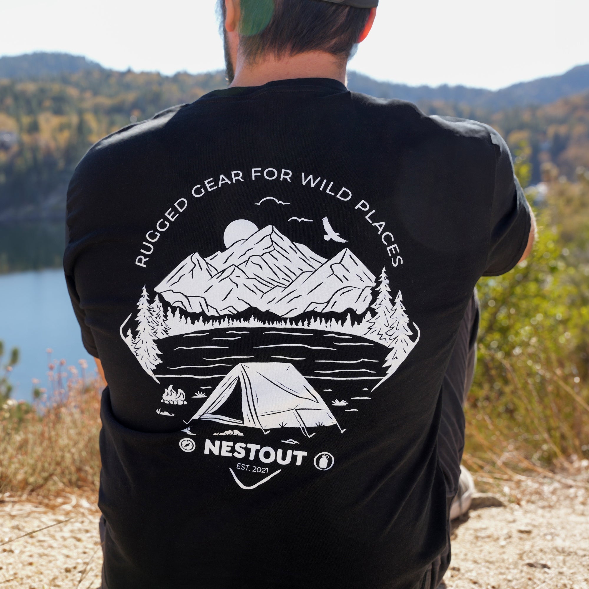Person wearing a black t-shirt with outdoor design and Nestout logo, standing in a scenic location.