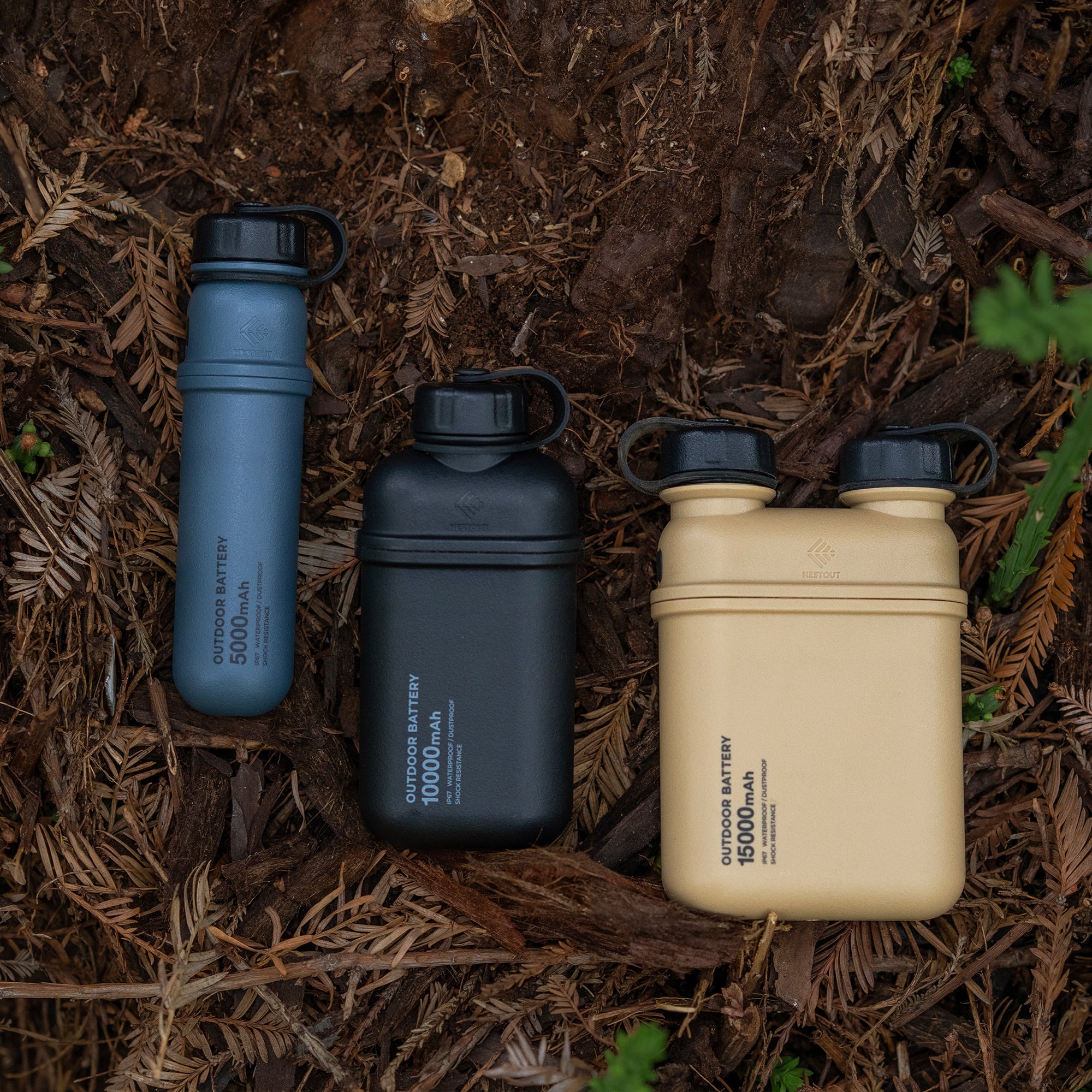 Image of nestout 10k black 5k grey 15k beige colored cylinder battery packs resting on ground in the woods | features military IP67 rating | stores up to 10000N of power | rugged waterproof design | compact size | versatile charging | to be used with NESTOUT solar panels, fans, lamps, and flashes