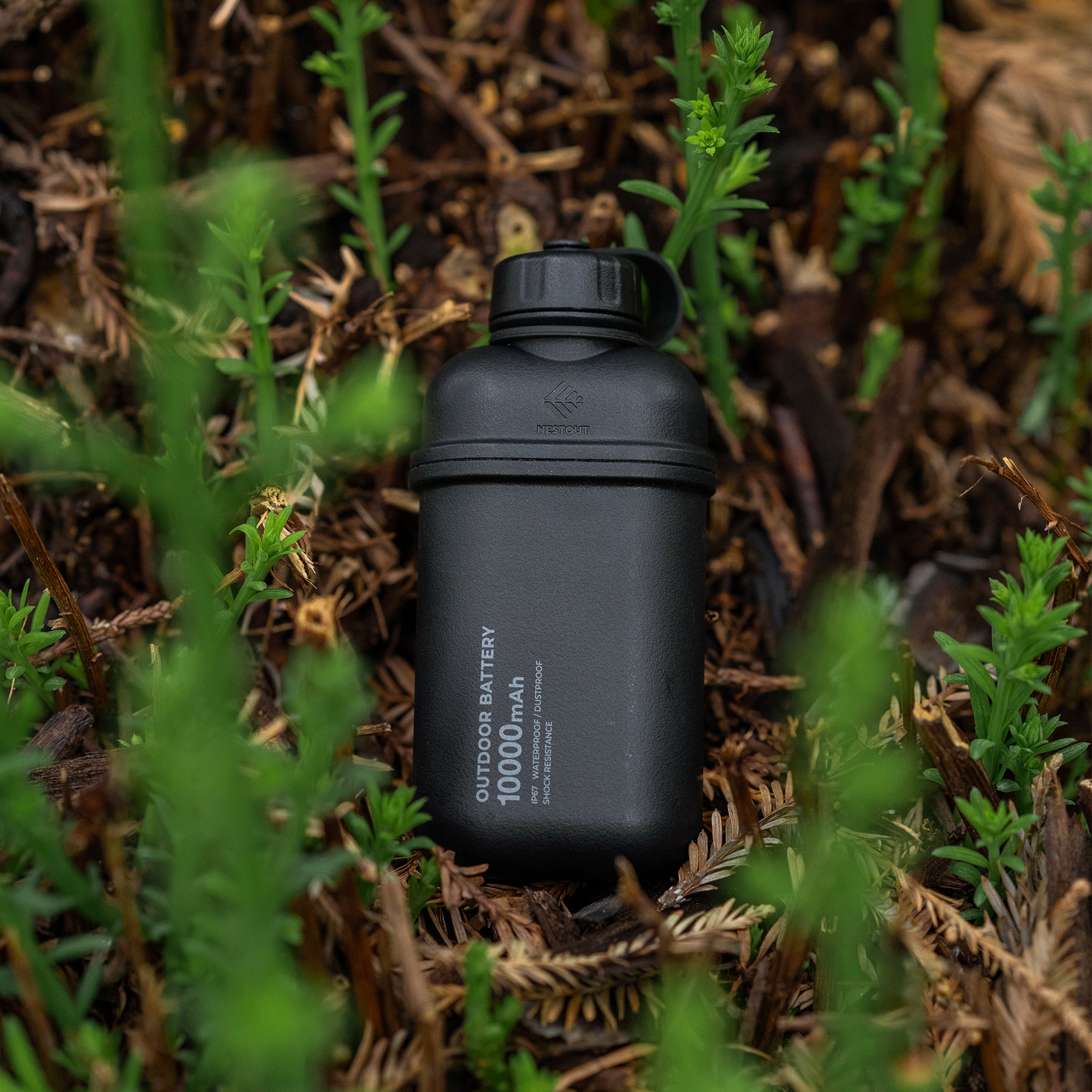 Image of nestout 10k black colored cylinder battery pack resting on shrubs | features military IP67 rating | stores up to 10000N of power | rugged waterproof design | compact size | versatile charging | to be used with NESTOUT solar panels, fans, lamps, and flashes