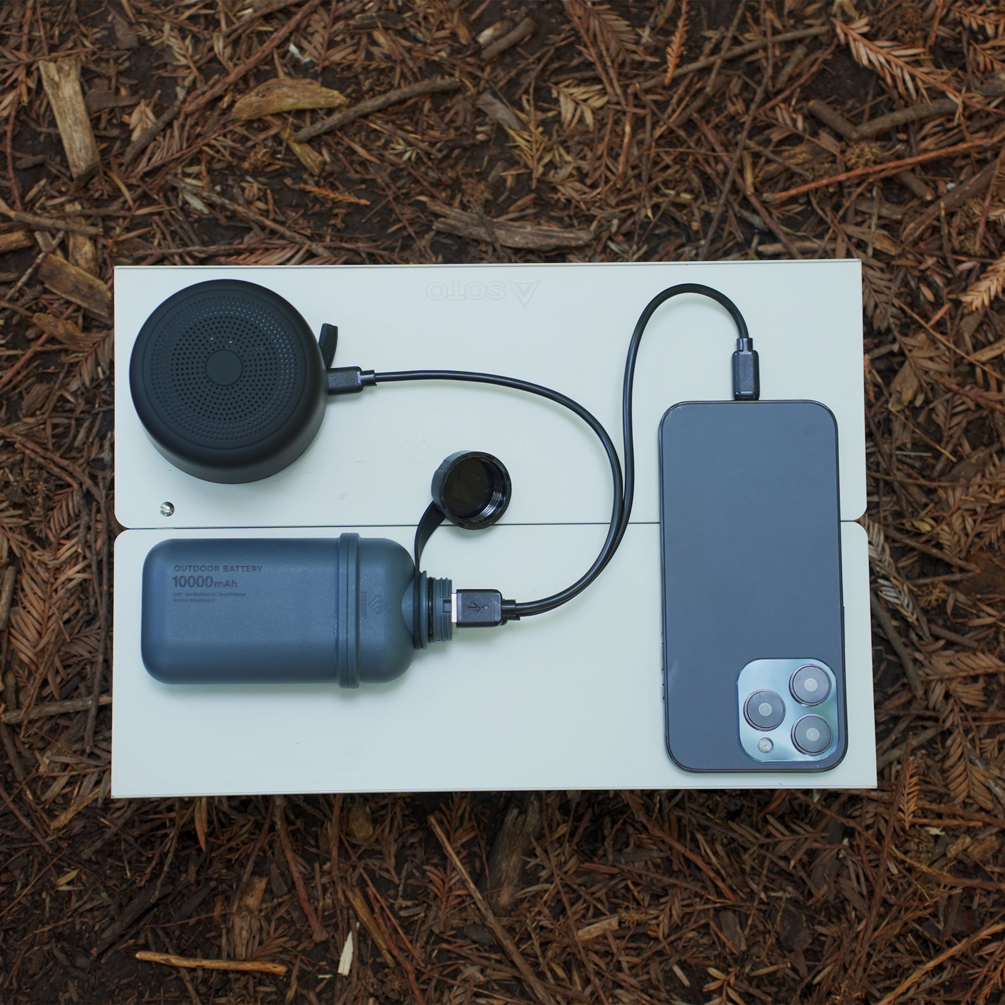 NESTOUT grey 10000mAh battery pack connected to smartphone and speaker laying on a small table in mulch