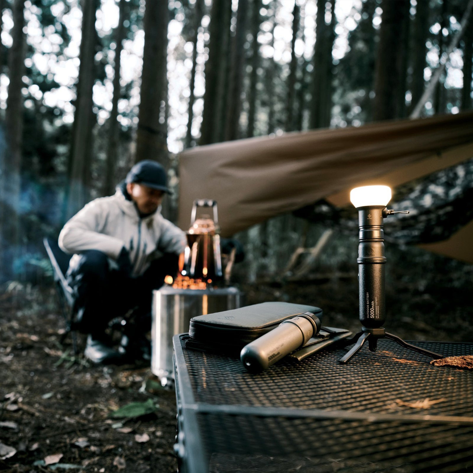 Image of nestout 5k black colored cylinder battery pack with nestout lamp connected standing on tripod on a table in the woods with person sitting around a camp boiling water | features military IP67 rating | stores up to 5000N of power | rugged waterproof design | compact size | versatile charging | to be used with NESTOUT solar panels, fans, lamps, and flashes