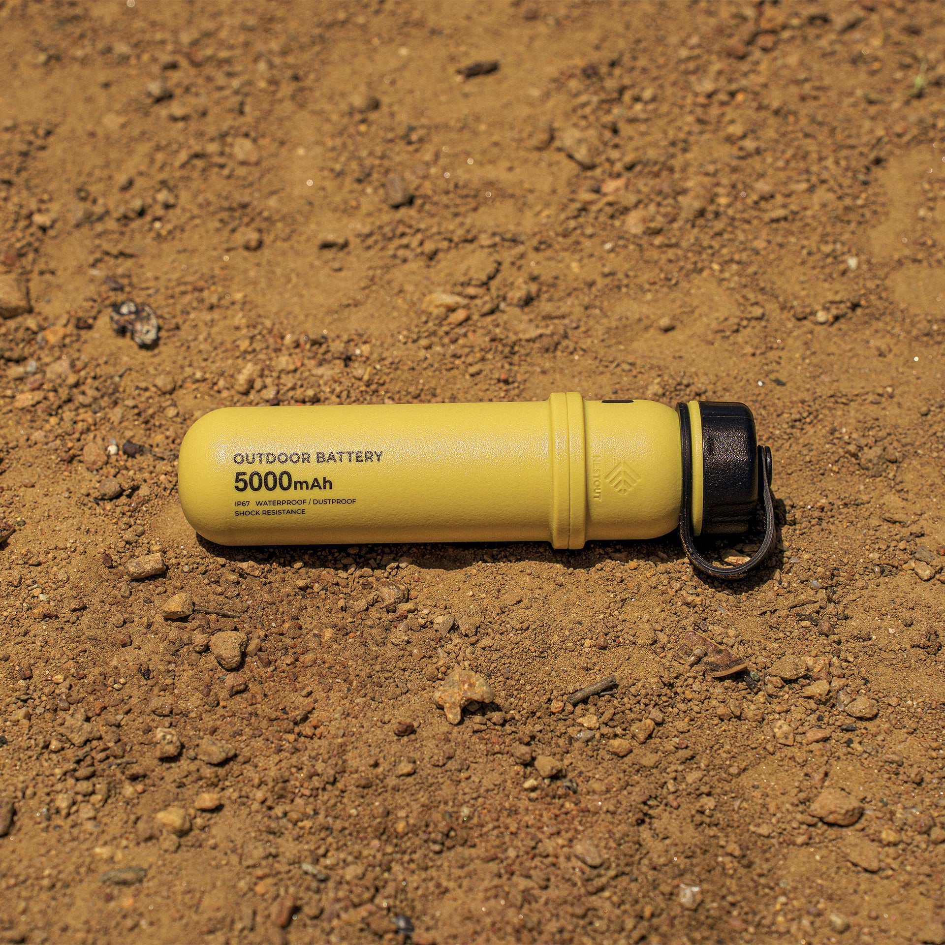 Mustard colored cylinder battery pack laying on dirt at Jenks Lake, showcasing the IP67 waterproof, dustproof, and shock resistance
